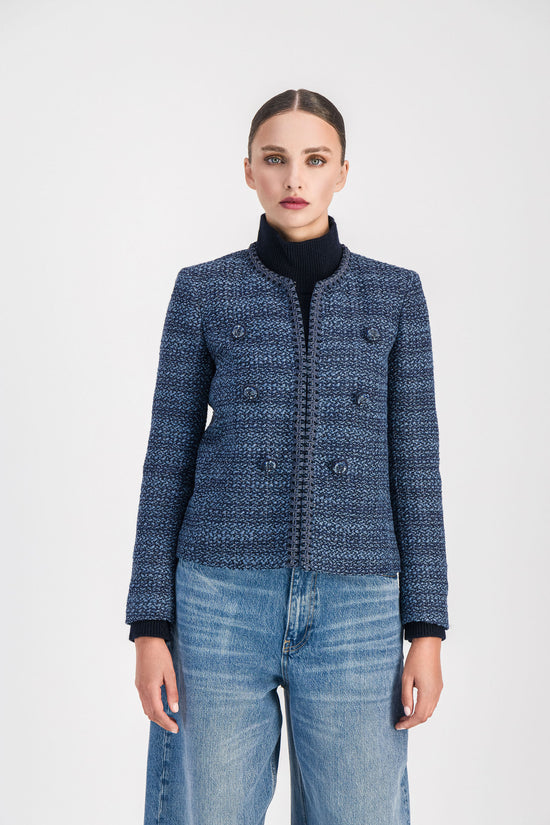 Gros plan sur un modèle portant une veste courte en tweed bleu denim présentant une coupe structurée, un col arrondi, des bordures tressées, un boutonnage apparent et des poches italiennes.