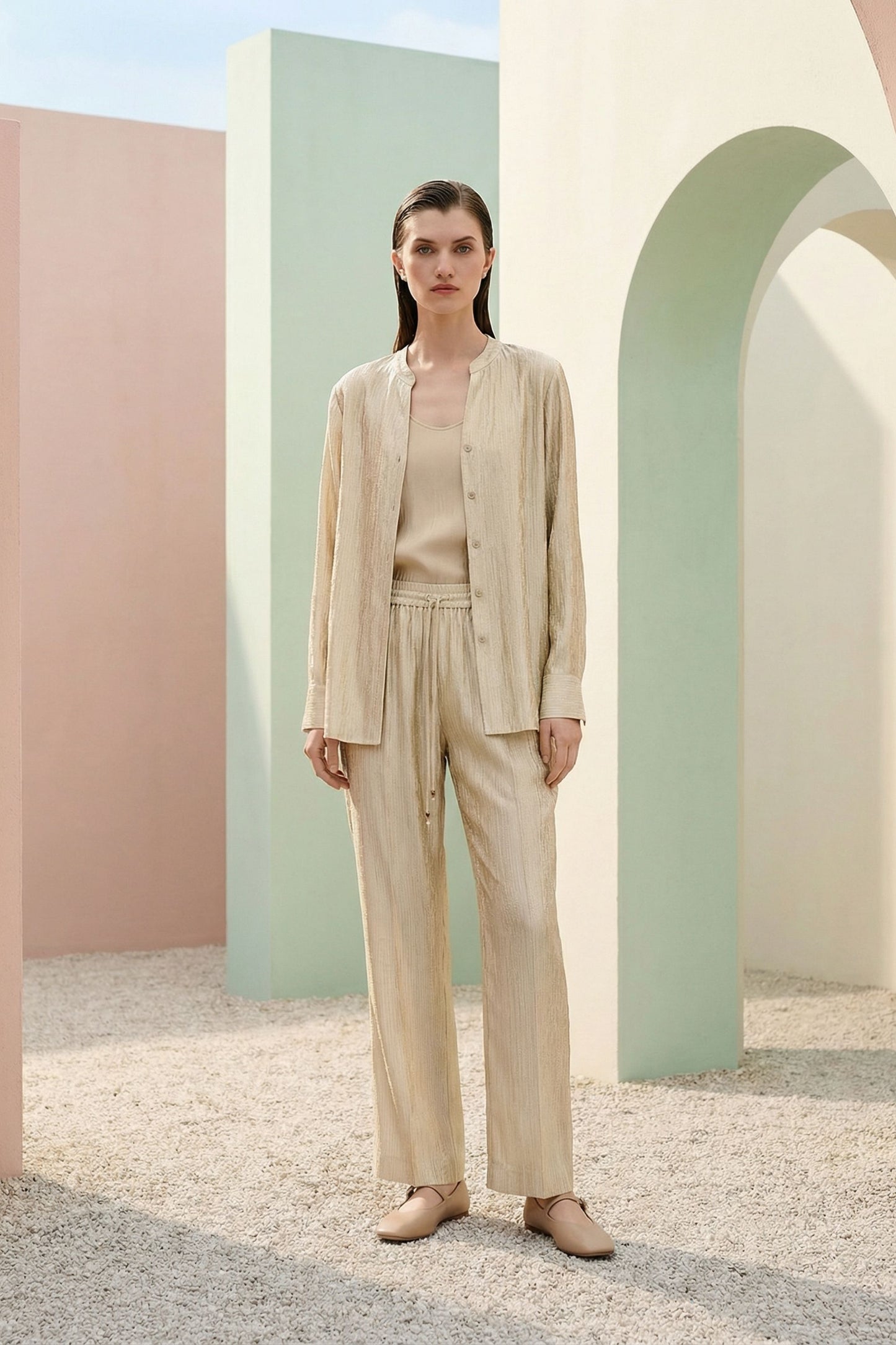Mannequin portant un ensemble beige composé d’une chemise légère et d’un pantalon droit à taille élastiquée, associé à des ballerines, dans un décor aux arches pastel.